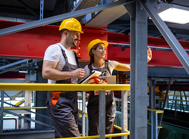 Hombre y mujer con casco amarillo y mono de trabajo, de pie sobre una plataforma elevada en una nave industrial